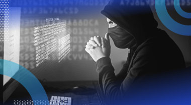 A stereotypical hacker sitting in front of a computer wearing a black hoodie and face mask, hands clasped in front of his face in concentration as he stares at holographic code that’s hovering off the screen.