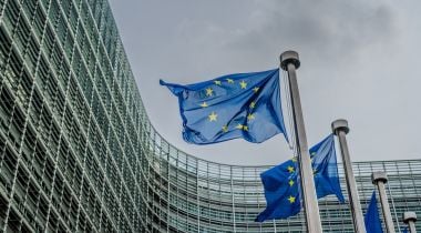 Two European Union flags wave in the wind, with a building surrounding them in the background.