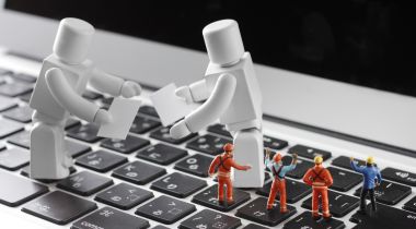 A photo of toy workers and two larger toy robots on a keyboard to show the idea of engineers building AI.