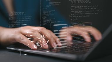 A close up of hands typing on a laptop with holographic images of code and AI icons floating in the foreground.