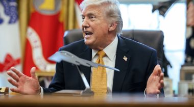 President Trump sits in the Oval Office wearing a dark suit and mustard-gold tie.