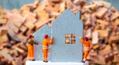 A photo of miniature figures building a small cut out of a house to show the idea of real estate tech developer teams working together. 