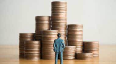 A miniature man in a business suit stands on a table looking at nine stacks of pennies.