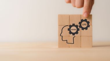 A photo of a hand placing wooden blocks with images of gears that say work and life next to a human head profile to show the idea of work-life balance and employee wellbeing. 