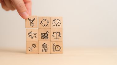 A photo of a hand with wooden blocks with physical, mental and emotional health icons to show the idea of employee wellbeing. 