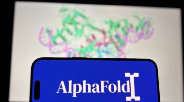 A mobile phone with the AlphaFold logo displayed on it, with an image of a protein structure on the wall behind it.