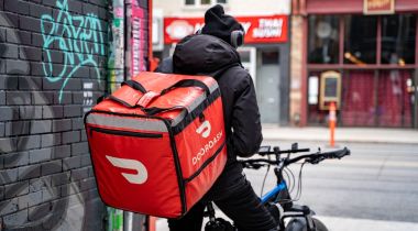 A DoorDash driver sits on a bike next to a wall.