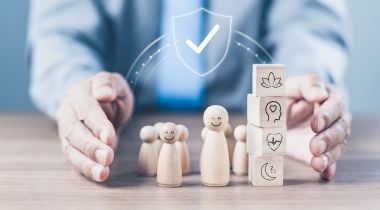 Hands around wooden figures with smiles and blocks stacked next to them with icons like plant, heartbeat pulse and a moon with sleep symbols to show the idea of protecting employee wellbeing. 