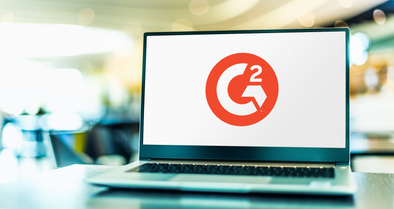 G2's red logo on a laptop. The laptop sits on a desk. A soft-focus office is visible in the background.