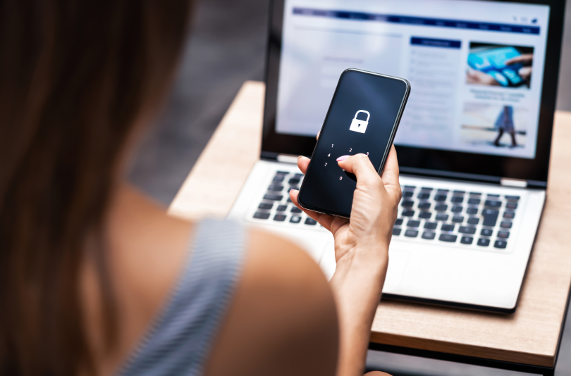 woman at computer looking at her phone which is prompting her for a code