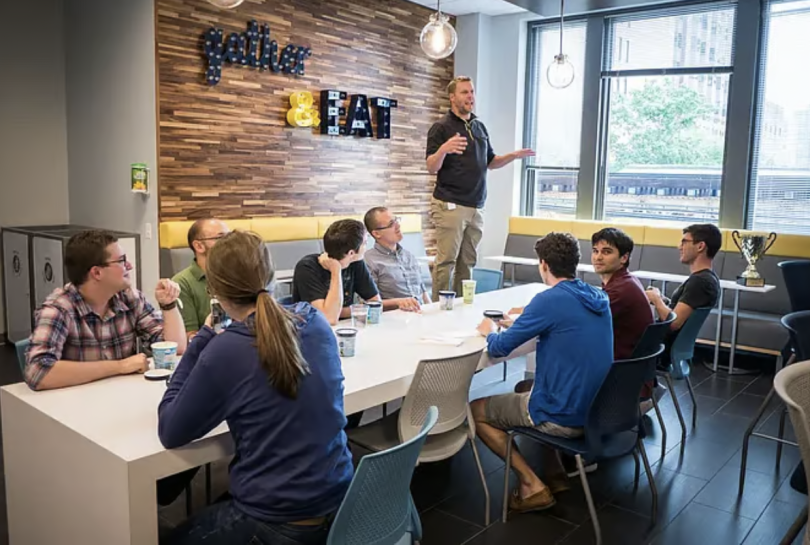Man standing on chair while eight people sit on chairs around conference table.