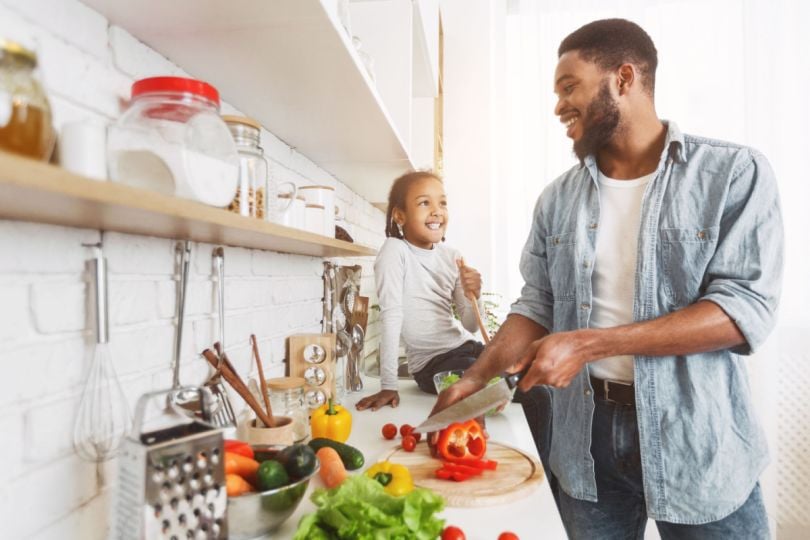 A father and daughter cheerfully prepare a meal together in a brightly lit kitchen. 