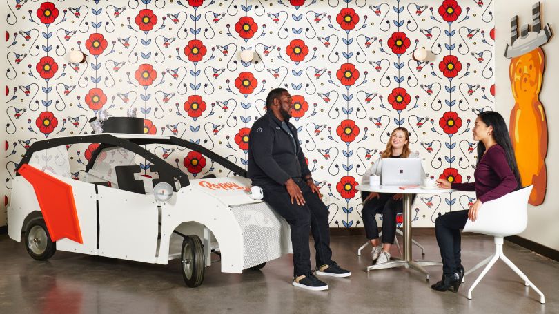 Three colleagues talk inside Poppy's Cafe in Cruise's headquarters; one is sitting on the hood of a vehicle model for Cruise's driverless cars