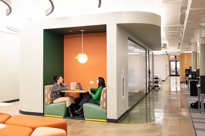 Two snap team members sit at booth-style meeting table in an office nook, meeting and talking with laptops open.