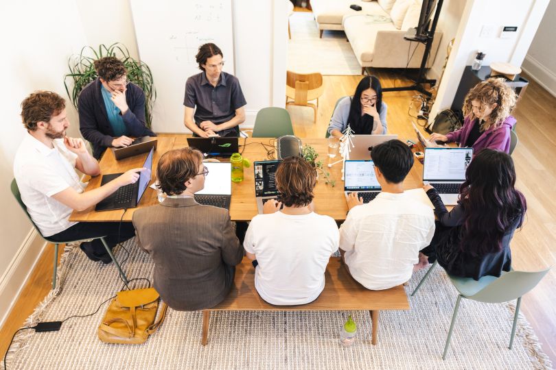 A group of people sit at a dining table working together on laptops. 