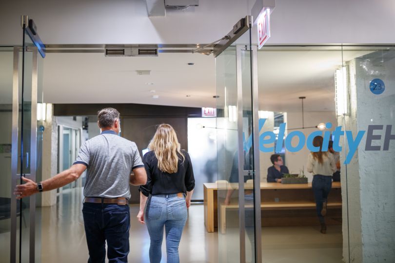 Candid photo of two team members entering the office through a glass door with a VelocityEHS logo on the glass wall next to it.