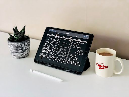 wireframes image of a tablet with a black screen and white sketchy wireframes. The tablet sits on a white desk with a green succulent and a white cup of coffee. There is a stylus on the desk.