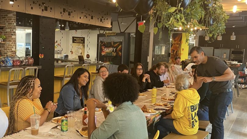 Sisense team members, a majority of them women, eat at a long dining table in the office kitchen space.