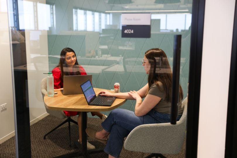 Two Zoro women team members meet in a small room behind a glass door with laptops in front of them.