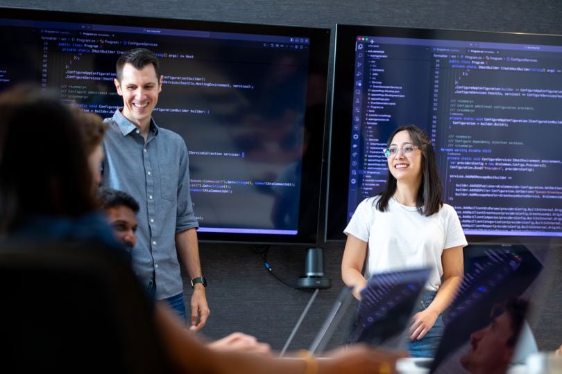 Two Built In engineers stand with code on the screens behind them, smiling at coworkers in front of them