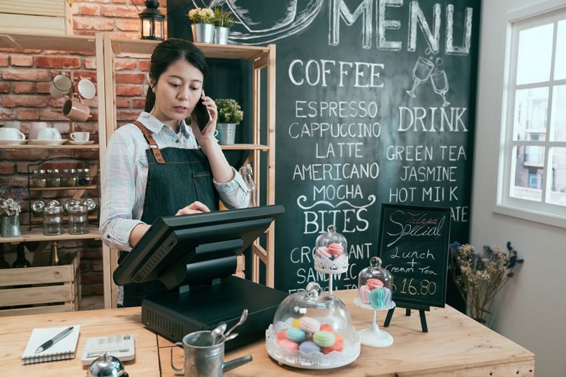  Female barista talking on mobile phone using digital tablet while standing behind counter in cafe