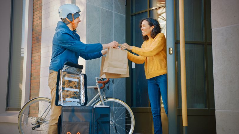 Food delivery man wearing thermal backpack on a bike delivers order to a female customer