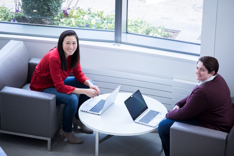 Photo of two women sitting with open laptops looking at camera smiling