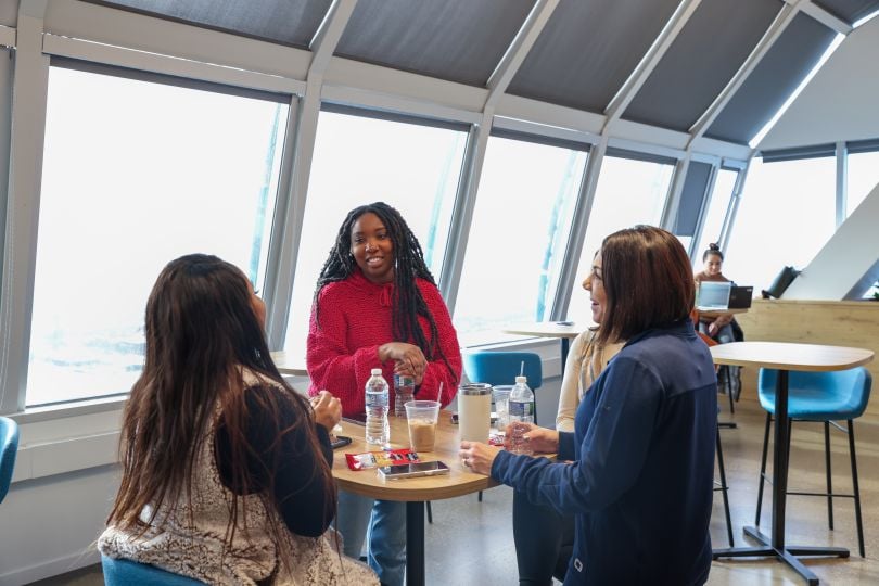 A group of four Zoro women team members taking a break together in a room full of windows, coffee and water bottles on the table in front of them. 