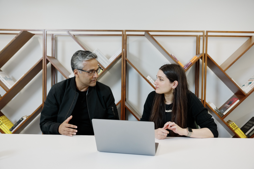 Two people sit at a table in front of an angled bookshelf, having a discussion. 