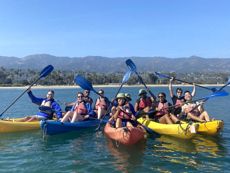 Alma team members gathered together in kayaks on lake, raising oars in the air