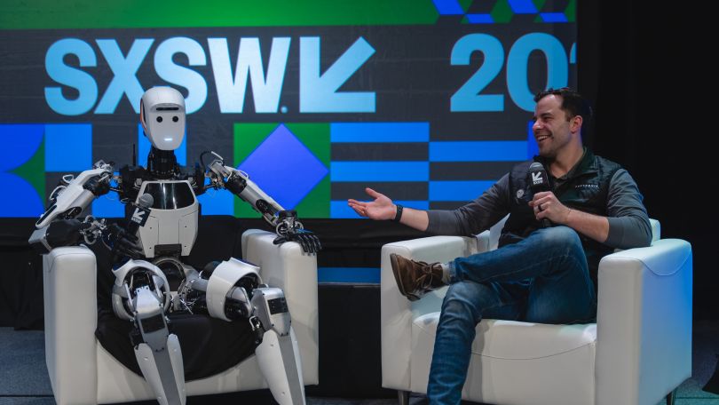 A leader from Apptronik sits on stage in a white chair with a humanoid robot, like an interview. They are in front of a sign saying SXSW in blue and green. 