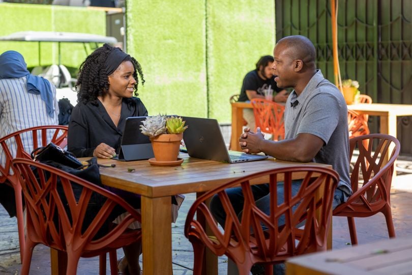 Photo of two NBCUniversal colleagues sitting at a table outside with laptops, talking and smiling