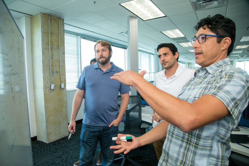 Three colleagues standing at whiteboard talking, gesturing.