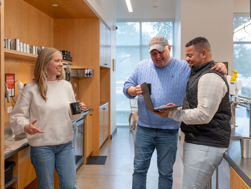 three employees in the break room with drinks and a laptop