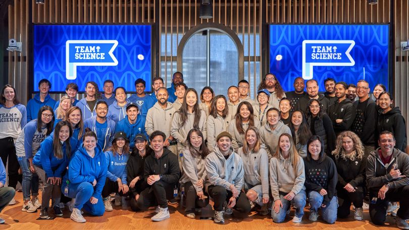  A group of Benchling employees stand for a group photo in front of two signs that say “Team Science.” 