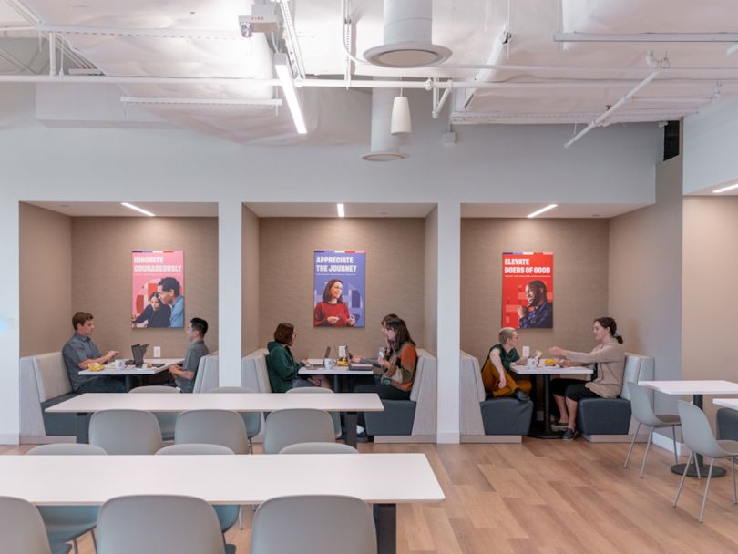 Three partitioned tables with employees sitting together in pairs at each. Behind them are three posters. One reads "Innovate Courageously," one reads "Appreciate the Journey" and one reads "Elevate the Doers of Good."