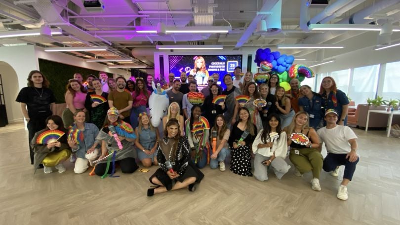 Pride@Braze ERG members gather in the office for an event, holding rainbow fans with balloons in the background.