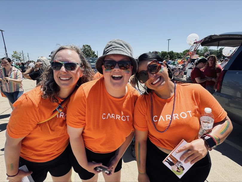 Three employees of Carrot Fertility wearing orange shirts and rainbow sunglasses smiling for a picture.