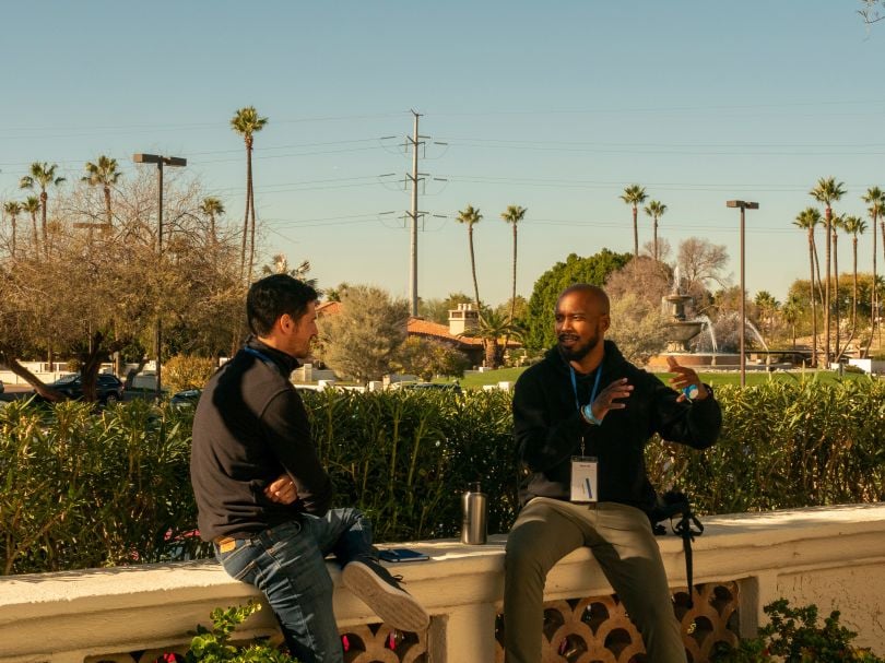 Two Chainlink Labs team members chat while sitting on an outdoor patio railing with palm trees in the background.