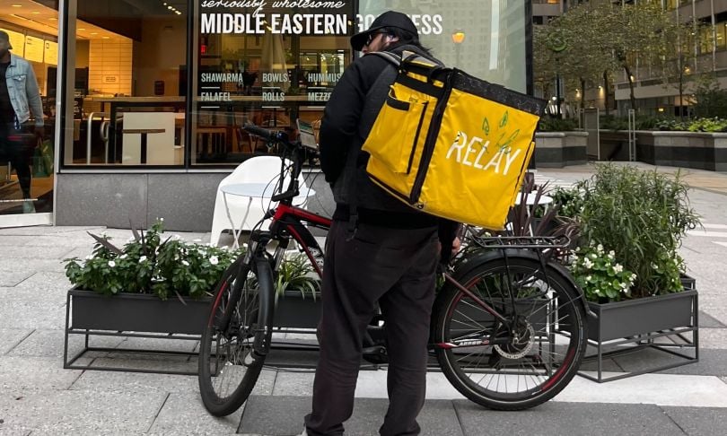 A Relay Delivery person stands outside restaurant with bike and cooler backpack.