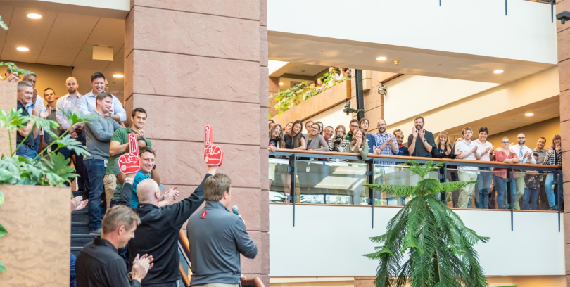 EchoStar team members stand along a balcony cheering as they watch leaders on a nearby staircase speak to them.