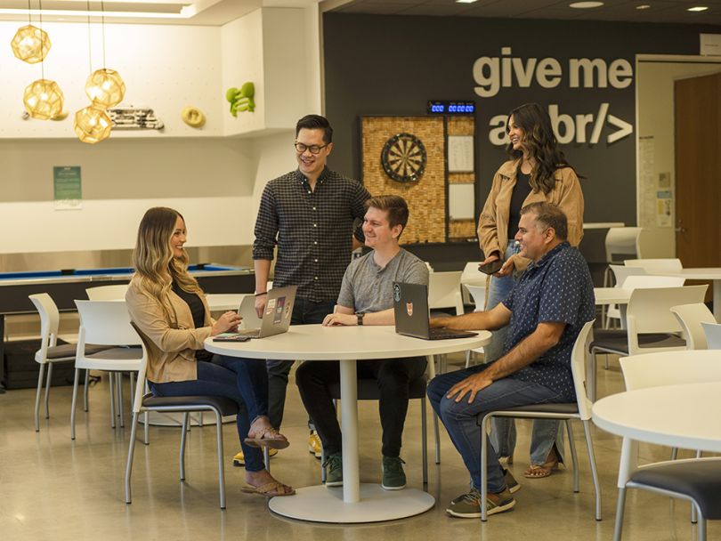 MobilityWare team members sit around a table in the breakroom, looking at laptops and talking with one another.