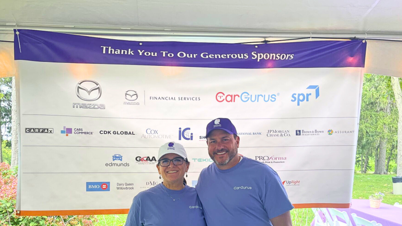 Two CarGurus team members stand in front of a banner showing sponsors at a dealer event.