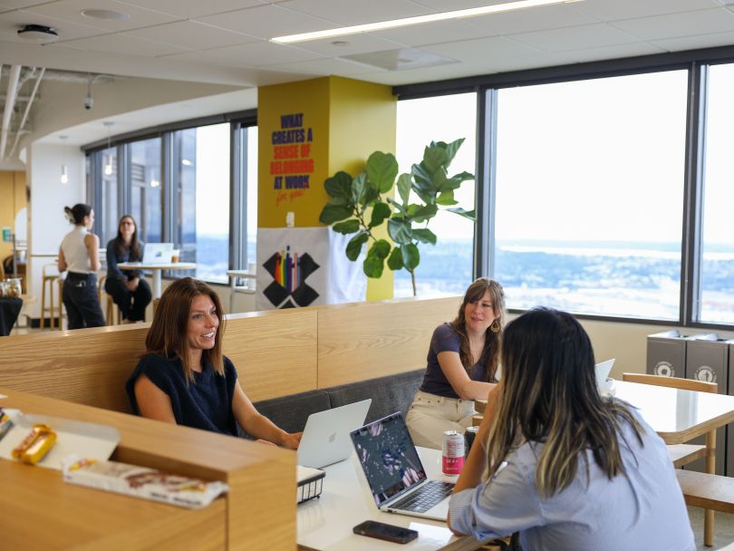 Dropbox team members seated with laptops chatting with each other in office next to window with aerial view of city