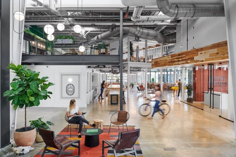 An open-concept two-level collaboration space is seen. It has an industrial yet homey vibe. A person walks through with a bike.