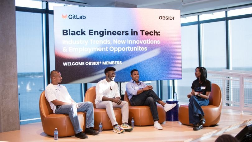 GitLab team member leads a panel discussion featuring Obsidi team members, all seated on a stage in front of windows looking out over a body of water. The screen behind the panel reads, “Black Engineers in Tech: Industry Trends, New Innovations and Employment Opportunities.”