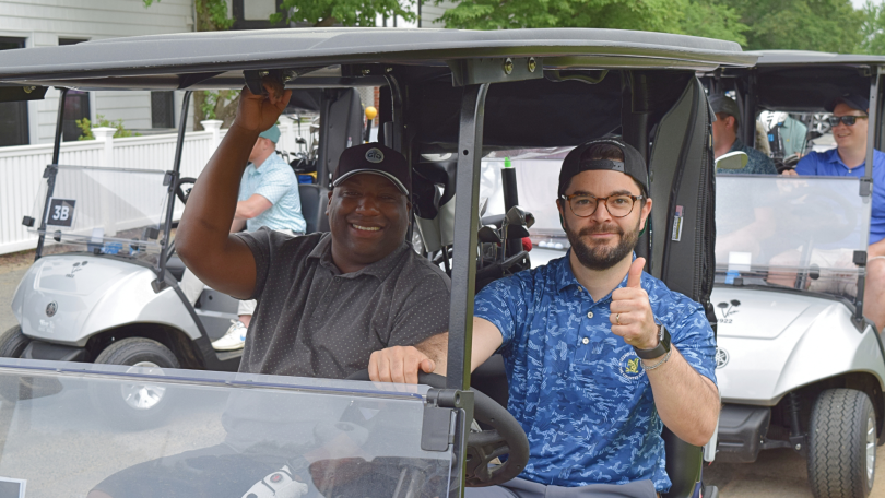 CarGurus team members smile and give a thumbs-up while driving a golf cart at a charity event. 