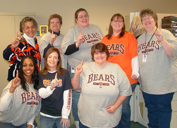 Group photo of Applied Systems colleagues, including Heather Prince in the back row, wearing Chicago Bears shirts and holding up their pointer finger to say “number one.”