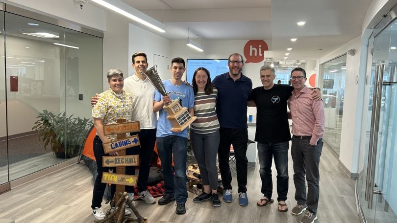 A photo of Hi Marley employees standing in a workplace hallway, holding a trophy and a wooden sign that reads “cabin,” “workshop,” “rec hall” and “tech cafe.”