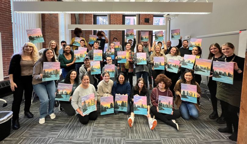 Members of Robin's team posing for a photo, each holding their own version of a painting on canvas of a sunset and pine trees.